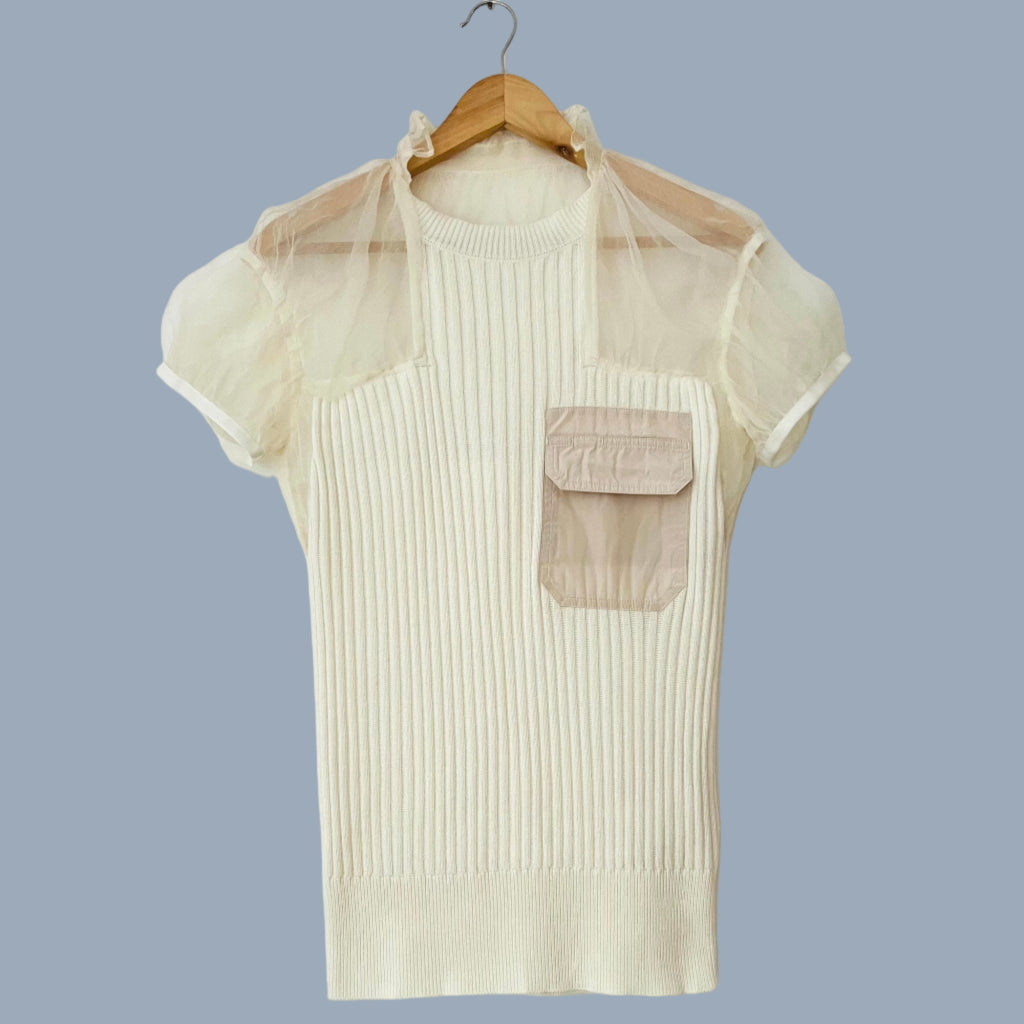 A cream-colored Sacai silk/cotton knit top with a high neckline, front contrasting pocket, and organza silk sleeves displayed on a hanger against a blue background.